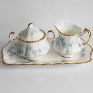 May include: A white porcelain tea set with a sugar bowl, creamer, and serving tray. The set is decorated with blue floral patterns and gold trim. The sugar bowl has a lid and handles. The creamer has a spout and handle. The tray has a scalloped edge.