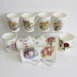 Könnte beinhalten: Eine Sammlung von Keramikbechern mit verschiedenen Blumen- und Landschaftsdesigns. Die Becher haben goldene Ränder und weiße Griffe. Einige Becher zeigen farbenfrohe Blumenmuster, andere malerische Landschaftsillustrationen.