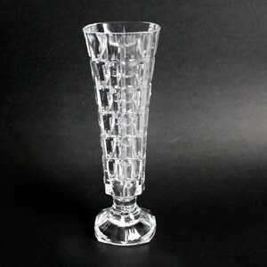 May include: A clear, faceted glass vase with a flared top and a pedestal base. The vase features a geometric, block-like pattern. The base is octagonal. The vase is set against a dark background.