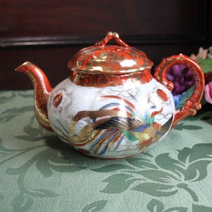 May include: A porcelain teapot featuring a white base with a vibrant orange and gold lid, handle, and spout. The body is decorated with a colourful phoenix motif, gold accents, and stylised cloud patterns in red, blue, and green over a white surface.