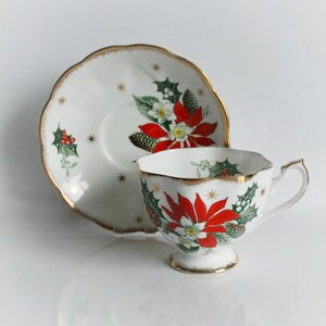 May include: A white teacup and saucer set with a gold trim and a festive poinsettia design. The cup and saucer feature red poinsettias, green holly, and gold star accents, ideal for Christmas. The set is a classic design.
