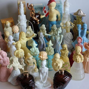 May include: A selection of vintage perfume bottles, each with a figurine topper in pastel shades. The figurines depict a variety of characters, including a boy with a Canadian flag and a woman skipping. The bottles are brown and clear.