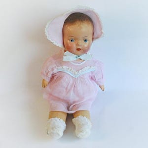 Puede incluir: Una muñeca de bebé vintage con una cara pintada, ojos azules y un vestido rosa. La muñeca lleva un gorro a juego con ribete de encaje y un lazo azul claro. Los calcetines blancos completan el conjunto. Un juguete coleccionable clásico.
