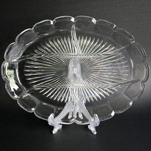 Puede incluir: Fuente de vidrio transparente con tres compartimentos y borde festoneado. La fuente ovalada presenta un diseño texturizado detallado que irradia desde el centro. Se exhibe sobre un soporte de plástico transparente, ideal para aperitivos.
