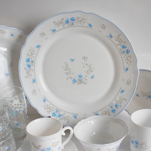 May include: A collection of white porcelain dinnerware with a blue floral pattern. The set includes plates, cups, bowls, and glasses. Each piece has a scalloped edge and a delicate blue floral design.