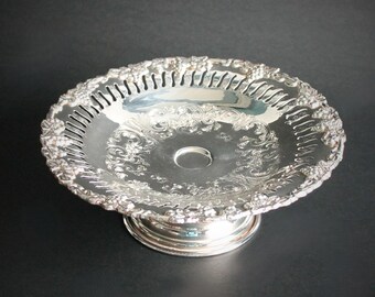 May include: A silver-colored, ornate pedestal bowl with a pierced rim and decorative floral and scrollwork patterns. The bowl has a round base and a central circular indentation. The bowl is likely made of metal.