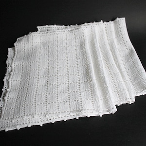 Könnte beinhalten: Ein Stapel weißer gehäkelter quadratischer Tücher. Die Tücher haben ein strukturiertes, offenes Webmuster und einen gewellten Rand. Die handgefertigten Tücher sind ordentlich gefaltet und auf einer dunklen Oberfläche angeordnet.