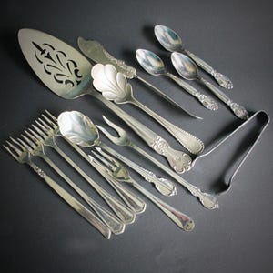 May include: A collection of antique silver serving utensils, including forks, spoons, a cake server, and a butter knife. The silverware features ornate floral and shell designs, with serving tongs. The pieces are arranged on a dark surface.