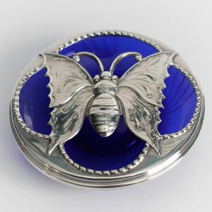 May include: A circular, silver-coloured container featuring a detailed butterfly design. The butterfly sits atop a deep blue glass base. The container has a beaded edge, likely a decorative item.