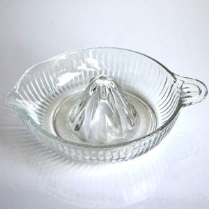 Peut inclure: Presse-agrumes en verre transparent avec un bol strié et un presse-agrumes central. Le presse-agrumes a une poignée et un bec verseur. Design simple et fonctionnel, adapté à la cuisine.