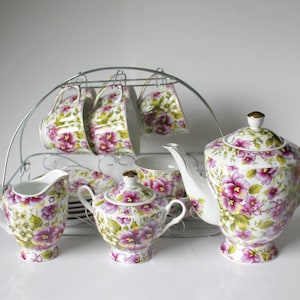 Może przedstawiać: Zestaw do herbaty w kwiatowym wzorze z czajnikiem, dzbankiem na śmietanę, cukiernicą i czterema filiżankami i spodkami. Zestaw jest ozdobiony kwiatowym wzorem w kolorze fioletowym i zielonym.