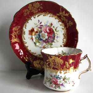 May include: A vintage teacup and saucer set. The set features a red and gold rim with floral designs. The teacup and saucer have a white center with a colorful floral bouquet. The set is displayed on a black stand.