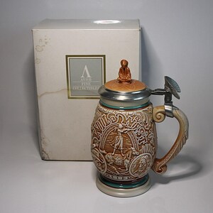 May include: A decorative beer stein with a detailed relief design, featuring a figure on horseback and other historical elements. The stein has a hinged lid, a handle, and a base. The stein is accompanied by a box with the text "AVON FINE COLLECTIBLES".