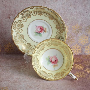 May include: A vintage teacup and saucer set with a pale yellow background and gold trim. The cup and saucer feature a delicate floral design with pink roses.