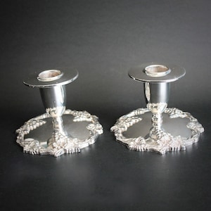 Könnte beinhalten: Zwei silberne Kerzenhalter mit klassischem Design. Jeder Halter hat einen kreisförmigen Sockel mit einem dekorativen Blumenmuster und eine zentrale Tasse zur Aufnahme einer Kerze. Die Kerzenhalter sind aus glänzendem Silbermetall gefertigt.