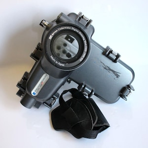 Puede incluir: Una carcasa gris Epoque Underwater Digital Housing Mod. EHS-1000 HD para cámara digital con una correa de muñeca negra. La carcasa de la cámara tiene una lente transparente y está etiquetada como "Made in Japan".
