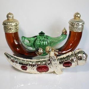 May include: Two curved amber glass bottles with gold-toned ornate caps, a green glass genie lamp, and a gold-toned decorative dagger-shaped bottle with red inset stones. These items are arranged together, showcasing distinct glass and metal textures.