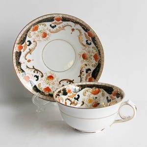 May include: A white teacup and saucer with a black, orange, and gold floral pattern. The pattern features stylized flowers and leaves.