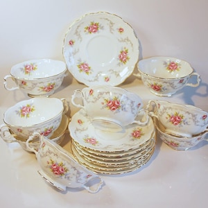 Puede incluir: Un juego de té vintage con tazas, platillos y una fuente de porcelana blanca. Cada pieza está adornada con motivos florales de rosas rosas y ribetes dorados. El juego incluye varias tazas, platillos y una fuente, perfecto para una merienda.