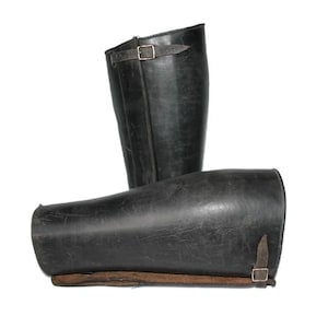 May include: A pair of black leather gaiters featuring a smooth, matte finish and a structured, tall design. Each gaiter includes a thin, adjustable leather strap with a small metal buckle for secure fastening, showing visible signs of wear.