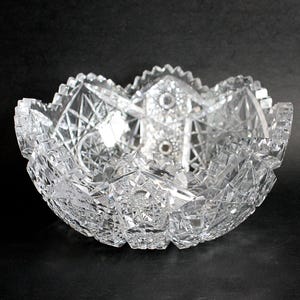May include: A clear, cut-glass bowl with a starburst pattern and a scalloped rim. The bowl features intricate geometric designs and a central, raised design. The bowl is likely made of lead crystal and is displayed against a black background.