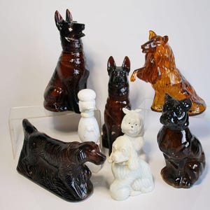 May include: A selection of ceramic dog ornaments in diverse breeds and colours. The figurines include a German Shepherd, a Collie, a Poodle, and a Bulldog. The colours vary from brown and black to white. The dogs are in various poses.