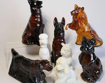 May include: A collection of ceramic dog figurines in various breeds and colors. The figurines include a German Shepherd, a Collie, a Poodle, and a Bulldog. The colors range from brown and black to white. The dogs are posed in different positions.