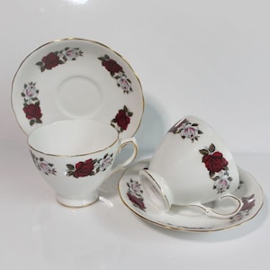 May include: Two white porcelain teacups and two matching saucers featuring a floral pattern of deep red and pale pink roses with green leaves. Each piece is finished with a thin gold trim along the scalloped edges and the cup handles.