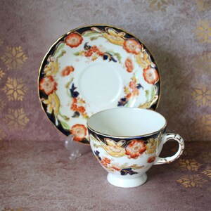 Può includere: Un set vintage di tazza da tè e piattino con un motivo floreale in tonalità di blu, arancio e giallo. La tazza e il piattino hanno un bordo dorato.
