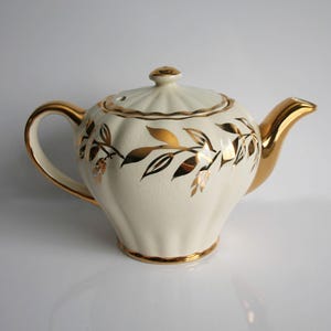 May include: Cream-colored ceramic teapot with gold accents. The teapot features a gold handle, spout, and rim. Gold leaf and branch designs adorn the body. The lid is cream-colored with a gold knob. A classic tea accessory.
