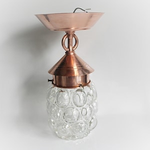 May include: Vintage copper-toned pendant light with a clear glass globe. The light fixture has a round, bubble-like glass shade and a copper-colored canopy and hardware. The fixture is designed to be mounted on a ceiling.