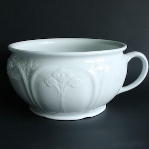 May include: A white porcelain bowl with a handle and floral relief detailing. The bowl has a wide rim and a rounded base. The handle is curved and attached to the side of the bowl. The floral relief is centered on the bowl's exterior.