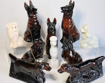 May include: A collection of ceramic dog figurines in various breeds and poses. The figurines are primarily brown and white, with some featuring glossy finishes. The dogs are arranged on a clear display surface, showcasing their detailed craftsmanship.