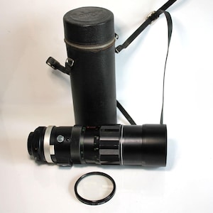 May include: A black camera lens with silver accents, a circular filter, and a black leather case with a shoulder strap. The lens is lying next to the case. The case has the word "Soligor" printed on the top.