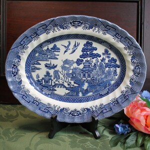 May include: An oval ceramic platter featuring a blue and white willow pattern. The design depicts a landscape with pagodas, trees, a bridge, and two birds in flight. The rim is decorated with a geometric border pattern in matching blue and white.