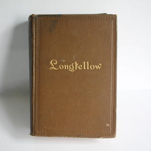 May include: A vintage book with a brown cover and the title "Longfellow" in gold lettering. The book has a textured appearance and a decorative border around the edges. The spine is dark brown.