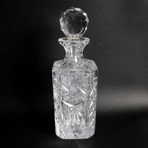 May include: A clear crystal decanter with a faceted stopper. The rectangular body features a starburst pattern, and the stopper is a large, multi-faceted sphere. The decanter is designed to hold liquids and is displayed against a black background.