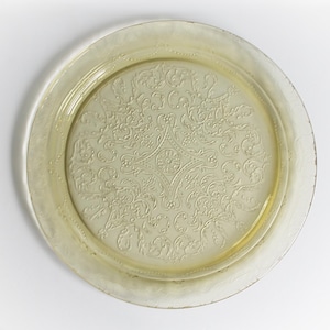 May include: A round, pale yellow glass tray with an embossed floral design. The tray has a slightly raised rim and a detailed pattern in the center. The overall design is symmetrical and elegant, suitable for serving or display.