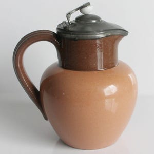 Può includere: Brocca vintage in ceramica con corpo arrotondato e manico marrone scuro. La parte superiore è di un marrone più scuro e ha un coperchio in metallo con un pomello bianco. La brocca è di colore pesca chiaro.