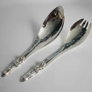 Puede incluir: Un par de cubiertos para ensalada plateados. Uno es una cuchara, el otro un tenedor. Ambos tienen una textura martillada en el cuenco y una figura detallada al final del mango.