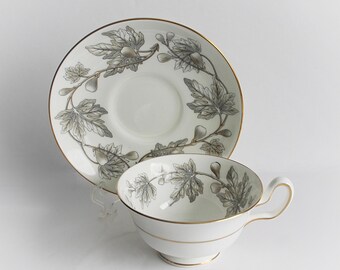 May include: A white teacup and saucer with a gold rim and a gray and white fig leaf design.