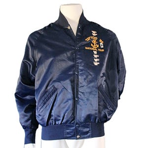 Op de afbeelding: Een donkerblauwe satijnen bomberjack met een gouden geborduurde anker en de tekst "Tattoo '85 National Tour".