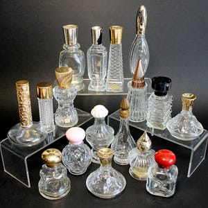 May include: A selection of vintage perfume bottles in diverse shapes and sizes, presented on clear acrylic risers against a black backdrop. The bottles have decorative stoppers in gold, silver, and black, with transparent glass bodies.