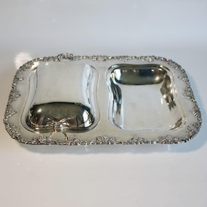 May include: A rectangular silver-toned serving tray featuring a polished finish and an ornate border with embossed grape and leaf motifs. The tray includes two recessed sections, one of which is covered by a removable domed lid with a small handle.