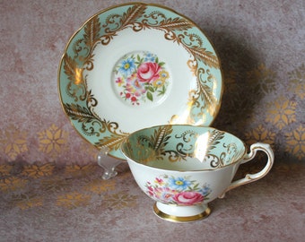 Peut inclure: Service à thé vintage en porcelaine fine. La tasse et la soucoupe sont blanches avec une bordure turquoise clair et des détails dorés. Le centre de la soucoupe et l'intérieur de la tasse présentent un motif floral avec des roses, des bleus et des jaunes. La base de la tasse est dorée.