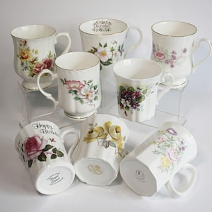 May include: A collection of white ceramic mugs with floral designs and gold trim. Some mugs feature roses, while others have various flower patterns. One mug has "Happy Birthday" text, and another has "December" text.