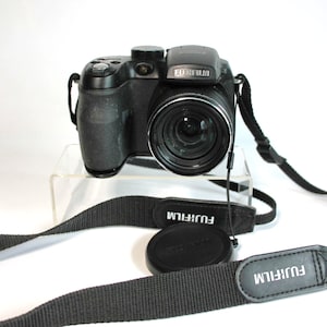 May include: A black Fujifilm digital bridge camera featuring a prominent lens, a textured grip, and a top-mounted flash unit. The camera includes a black neck strap with the Fujifilm logo printed in white and a separate circular black lens cap.