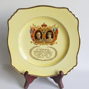 Op de afbeelding: Een vierkante, gele herdenkingsbord met een gouden rand. Het bord toont portretten van koning George VI en koningin Elizabeth, met tekst ter herdenking van hun bezoek aan Canada en de Verenigde Staten in 1939.