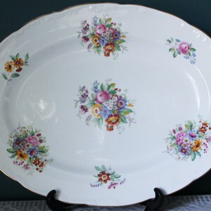 May include: An oval, white porcelain serving platter with a scalloped edge and gold trim. The platter is decorated with clusters of colourful flowers in shades of pink, blue, yellow, and orange. A decorative piece for serving food.