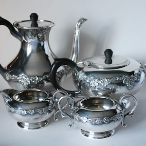 Puede incluir: Un juego de té de plata con tetera, jarra de crema, azucarero y un diseño a juego. El juego presenta un patrón floral y asas negras.